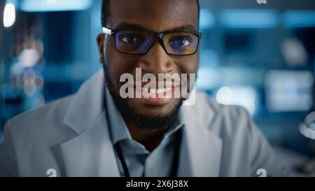 Schöner schwarzer Mann mit Brille, der charmant lächelt und die Kamera ansieht. Junger intelligenter männlicher Ingenieur oder Wissenschaftler, der im Labor arbeitet. Technischer Bokeh-Blauer Hintergrund. Nahaufnahme Stockfoto