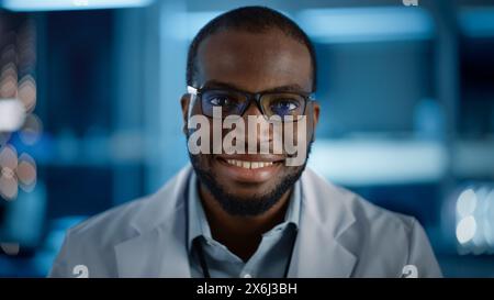 Schöner schwarzer Mann mit Brille, der charmant lächelt und die Kamera ansieht. Junger intelligenter männlicher Ingenieur oder Wissenschaftler, der im Labor arbeitet. Technischer Bokeh-Blauer Hintergrund. Nahaufnahme Stockfoto