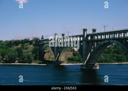 Eine alte Bogenbrücke überquert einen Fluss, mit Eisenbahngleisen oben und umgeben von üppigem Grün mit einem Hauch von städtischen Gebäuden im Hintergrund Stockfoto