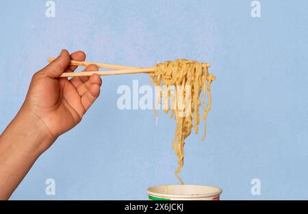 Hand mit Nudeln aus einer Instant-Suppe in einem Glas, gewürzte Nudeln auf hellblauem Hintergrund Stockfoto