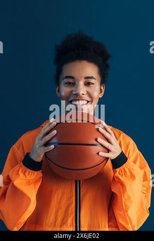 Porträt eines jungen lächelnden Basketballspielers. Fröhliche Frau in orangefarbener Sportswear, die Basketball über blauem Hintergrund hält. Porträt eines jungen Lächelns Stockfoto
