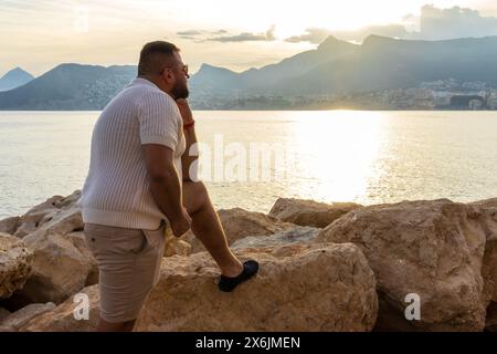 Dieses Porträt fängt einen 30-jährigen Mann ein, während er über die ruhige Weite des Mittelmeers reflektiert. Die untergehende Sonne wirft ein goldenes Leuchten über den Stockfoto