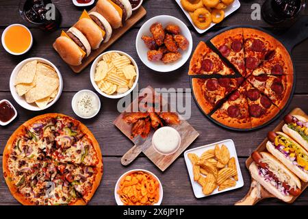 Junk Food Tischszene. Pizza, Hamburger, Hot Dogs, Chicken Wings und salzige Snacks. Blick von oben über dunklem Holzhintergrund. Stockfoto