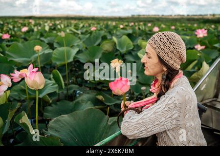 Eine Frau sitzt in einem Boot auf einem Feld rosa Lotusblüten. Sie trägt einen Hut und einen Schal. Stockfoto