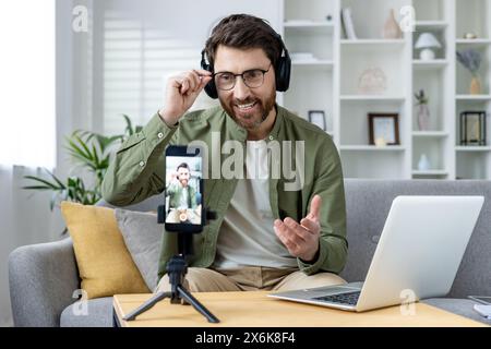 Lächelnder Mann mit Kopfhörern, der mit seinem Laptop und Smartphone online Videoanrufe führt, während er zu Hause auf dem Sofa sitzt. Stockfoto