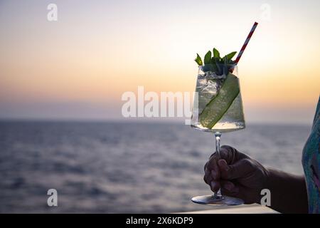 Hand mit Gin Tonic Cocktail auf dem Geländer des Expeditionskreuzfahrtschiffes SH Diana (Swan Hellenic) im Roten Meer bei Sonnenuntergang, auf See, in der Nähe von Saudi Stockfoto