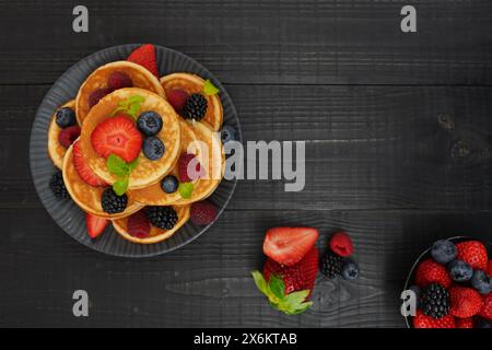 Ein flacher goldener Pfannkuchen auf schwarzem Holzhintergrund mit frischen Beeren sorgt für eine gemütliche und verwöhnende Szene. Stockfoto