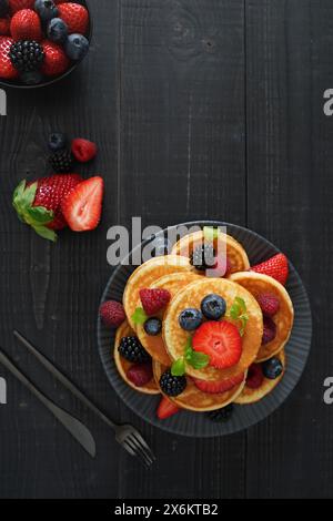 Ein flacher goldener Pfannkuchen auf schwarzem Holzhintergrund mit frischen Beeren sorgt für eine gemütliche und verwöhnende Szene. Stockfoto