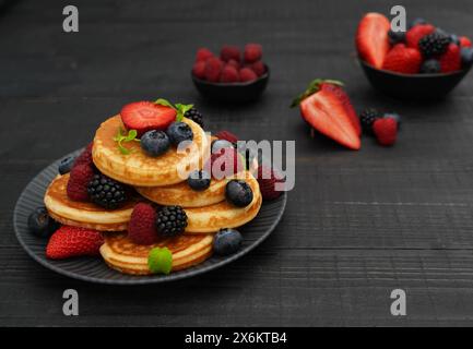 Nahaufnahme von gestapelten Pfannkuchen, die reich mit verschiedenen Beeren und frischen Minzblättern auf dunkler Holzoberfläche dekoriert sind Stockfoto