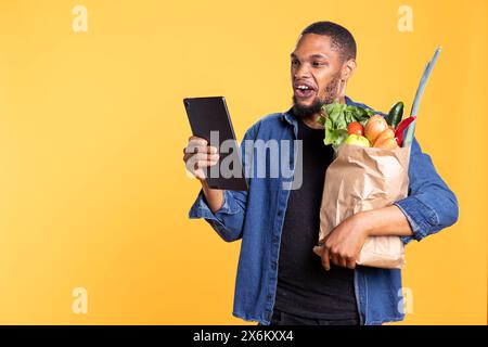 Ein afroamerikanischer Mann checkt seine Social-Media-Texte im Studio und trägt eine Papiertüte voller Obst und Gemüse aus dem Supermarkt. Selbstbewusster, entspannter Kerl, der Spaß beim Surfen im Internet hat. Stockfoto