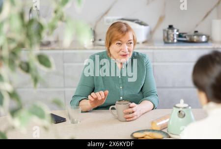 In der Küche am Tisch kommuniziert eine ältere lächelnde Frau mit einem Freund und trinkt Kaffee oder Tee Stockfoto