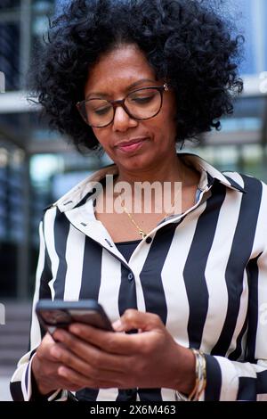 Erwachsene afroamerikanische Frau in formellem Anzug, die vor dem Bürogebäude mit Mobiltelefon steht Stockfoto