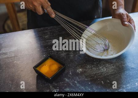 Bakers Hände schlagen Eier mit einem Handrührgerät auf einer weißen Platte, wobei Eigelb von Weißen getrennt wird. Kochen auf traditionelle Weise Stockfoto