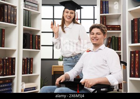 Männlicher Absolvent im Rollstuhl mit seinem Klassenkameraden in der Bibliothek Stockfoto