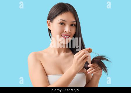 Junge asiatische Frau, die Haarserum auf blauem Hintergrund aufträgt, Nahaufnahme Stockfoto