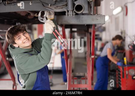 Lächelnder junger Mechaniker repariert Fahrgestell eines Autos im Aufzug in der Werkstatt Stockfoto