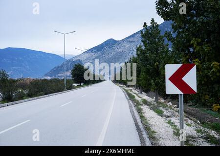 Leere Straße, die in Richtung eines Berges führt, mit einem roten Richtungsschild an der Seite. Stockfoto