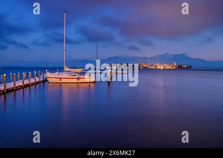 Schneebedeckter Bootssteg mit beleuchtetem Segelboot auf dem Chiemsee mit Fraueninsel- und Chiemgauer Alpen im Hintergrund, Chiemsee, Chiemgauer Alpen, Oberer BAV Stockfoto
