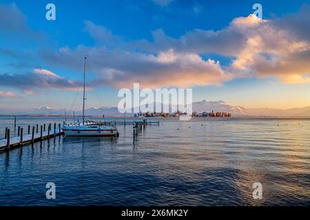 Schneebedeckter Bootssteg mit Segelboot auf dem Chiemsee mit Fraueninsel- und Chiemgauer Alpen im Hintergrund, Chiemsee, Chiemgauer Alpen, Oberbayern, Ba Stockfoto