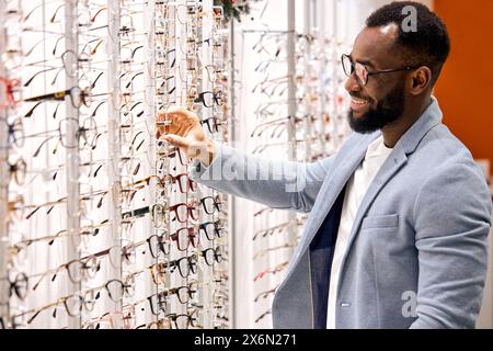 Hübscher junger, lächelnder bärtiger afrikanischer Mann, der eine Brille an der Optik kauft. Konsum, Shopping, Lifestyle, Medizinkonzept, Nahaufnahme von der Seite, Stockfoto