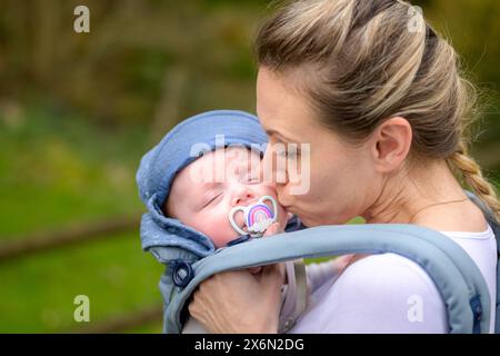 Glückliche Frau küsst ihr Baby, während sie es in einer Babytrage im Park hält und trägt Stockfoto