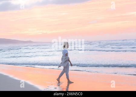 Eine ruhige Szene, während ein Individuum entlang der Küste spaziert und sich während eines lebendigen Sonnenuntergangs auf dem nassen Sand mit sanften Meereswellen reflektiert, die sanft abebben und fließen Stockfoto