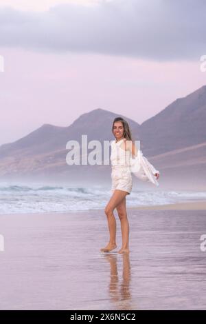 Glückliche Frau spaziert an einem ruhigen Strand mit Bergen im Hintergrund bei Sonnenuntergang, barfuß mit einer weißen Jacke über dem Arm und genießt den Frieden Stockfoto