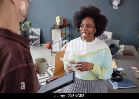 Porträt einer lächelnden Afroamerikanerin, die mit einem männlichen Kollegen plaudert, der während der Kaffeepause im Büro steht und eine Tasse hält Stockfoto