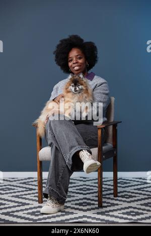 Vertikales Porträt in voller Länge einer lächelnden Afroamerikanerin, die einen Hund im Schoß hält und zu Hause in die Kamera schaut Stockfoto