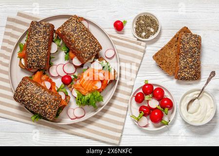 Räucherlachs-Sandwiches auf Roggenbrötchen mit frischem roten Rettich, Salat und Sahne frisch auf Teller auf weißem Holztisch mit Zutaten, horizontal Stockfoto