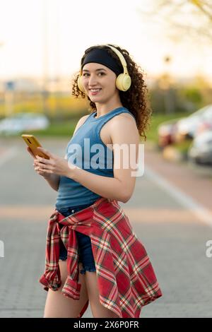 Lächelnde Frau mit lockigen Haaren, die ihr Smartphone benutzt und Kopfhörer trägt, in einem blauen Tanktop in einem pulsierenden Stadtpark bei Sonnenuntergang. Stockfoto