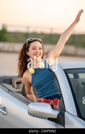 Überschwängliche junge Frau mit lockigen Haaren weht fröhlich aus einem Cabrio, mit Kopfhörern und Sonnenbrillen. Stockfoto