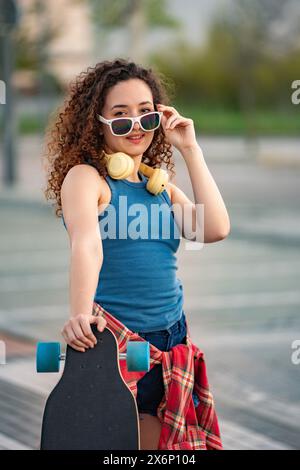 Modische junge Frau mit lockigen Haaren, die ihre Sonnenbrille anpasst, ein Skateboard hält und Kopfhörer in einem urbanen Park trägt. Stockfoto