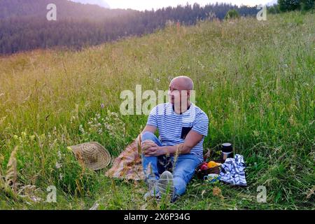 Reifer Mann, Senior sitzt am sanften Hang des Berges, neben Picknickkorb mit Obst, Thermos, grünem Gras auf der Wiese, Konzept des Familienpicknicks auf nat Stockfoto