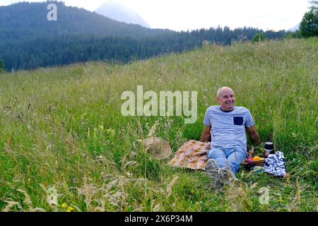 Reifer Mann, Senior sitzt am sanften Hang des Berges, neben Picknickkorb mit Obst, Thermos, grünem Gras auf der Wiese, Konzept des Familienpicknicks auf nat Stockfoto