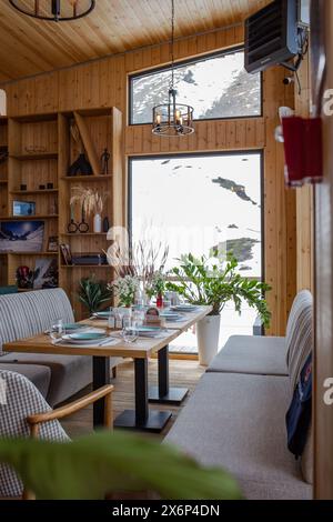 Ein rustikales Restaurant mit Holzvertäfelung, großen Fenstern, Blick auf die schneebedeckte Landschaft, Tischen, Stühlen, Kamin und Bar schaffen ein warmes Ambiente. Stockfoto