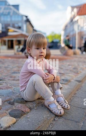 Porträt eines kleinen Mädchens. Ein wunderschönes, durchdachtes Mädchen, das 2-3 Jahre alt ist und legere Kleidung trägt, sitzt auf den Pflastersteinen auf einer europäischen Straße. Stockfoto