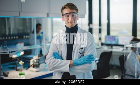 Medical Science Laboratory: Gutaussehender junger Wissenschaftler mit weißem Mantel und Schutzbrille, posierend und lächelnd, der die Kamera mit Kreuzen ansieht. Vielseitiges Team von Spezialisten. Mittlere Porträtaufnahme. Stockfoto