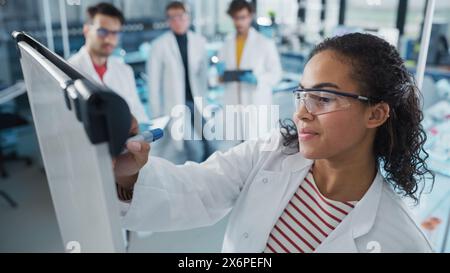 Medical Science Laboratory: Beautiful Black Female Scientist schreibt detaillierte Projektdatenanalyse auf den Vorstand, ihr vielfältiges Team von Kollegen hört zu. Nachwuchswissenschaftler Lösen Probleme. Stockfoto