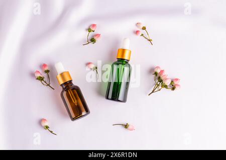 Zwei verschiedene Flaschen mit einem Tropfer mit Serum oder einem kosmetischen natürlichen Gesichts- und Körperpflegeprodukt auf weißem Satin unter dekorativen Beeren. Draufsicht Stockfoto