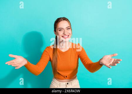 Foto eines freundlichen fröhlichen Mädchens mit Ingwerfrisur gekleidetem orangefarbenen Hemd, das die Hände spannt, um Sie isoliert auf blaublauem Hintergrund zu umarmen Stockfoto