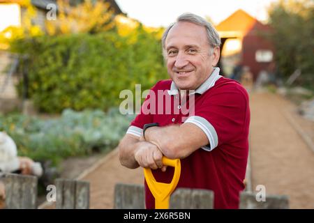 Ein erwachsener Mann stand entspannt neben dem Zaun, mit einer Schaufel im Garten, die lächelte Stockfoto