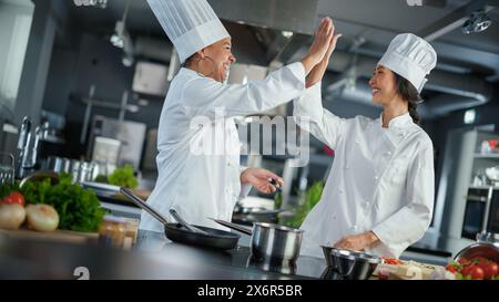 Restaurant Kitchen: Porträt asiatischer und schwarzer weiblicher Köche, die Gerichte zubereiten, Essen verkosten, High-Five in erfolgreicher Feier machen. Zwei Profis Kochen Köstlich, Authentisch Essen, Gesunde Mahlzeiten Stockfoto
