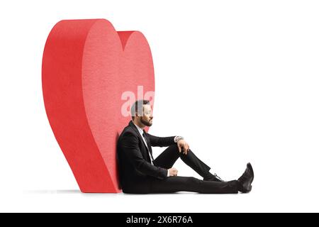 Nachdenklicher Mann auf Liebe sitzt auf dem Boden und lehnt sich auf ein rotes Herz isoliert auf weißem Hintergrund Stockfoto