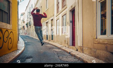 Positiver und glücklicher junger Erwachsener in legeren Kleidern tanzt aktiv, während er auf der Straße einer Altstadt in einer Stadt spaziert. Szene in einer urbanen ruhigen Narrow Town Street. Aufnahme von hinten. Stockfoto