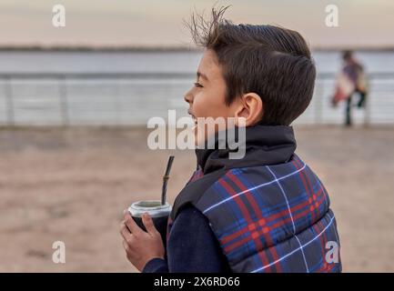 Close-up-Porträt eines latino-Jungen im Profil eines trinkenden Gefährten, der an einem Winternachmittag am Fluss in Argentinien lächelt. Stockfoto