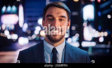 Porträt eines gutaussehenden bärtigen Mannes, der lächelnd in die Kamera schaut, in der Nachtstadt steht, mit Bokeh Neon Street Lights im Hintergrund. Glücklicher, Selbstbewusster Junger Mann Mit Stilvollem Anzug. Stockfoto