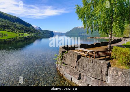 Ein ruhiger Blick auf den See mit einem hölzernen Picknicktisch unter einem üppigen Baum, umgeben von majestätischen Bergen und kristallklarem Wasser an einem hellen Frühlingsmorgen Stockfoto