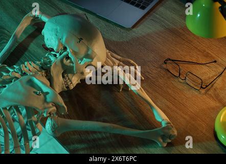 Wartendes Konzept. Menschliches Skelett, das an einem Holztisch mit Laptop schläft Stockfoto