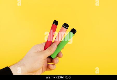 Elektronische Einweg-Zigaretten verschiedener Geschmacksrichtungen in der Hand auf einem gelben Hintergrund. Das Konzept von modernem Rauchen, Verdampfen und Nikotin. Stockfoto